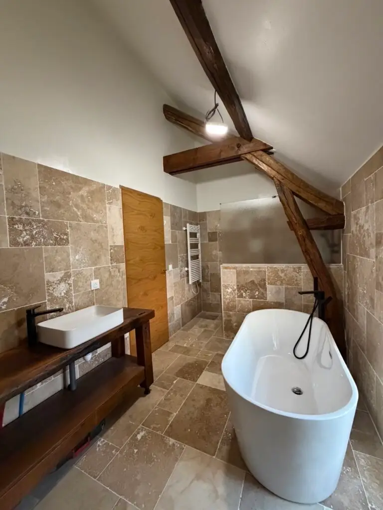 Salle de bain rénovée à Malesherbes : baignoire îlot, poutres apparentes et sol en pierre naturelle